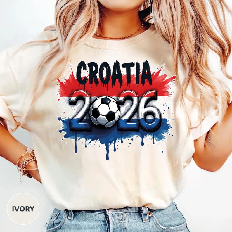 Może przedstawiać: Kremowa koszulka z czarnym napisem "CROATIA" nad liczbami "2026" i piłką nożną. Projekt wykorzystuje kolory czerwony, biały i niebieski z efektem rozprysku.