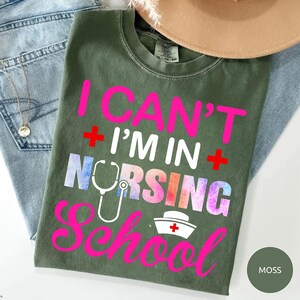May include: A moss green short-sleeve t-shirt featuring the hot pink and white text I CAN'T I'M IN NURSING SCHOOL with two red plus signs, a white stethoscope graphic replacing the letter U, and a white nurse cap icon with a red cross.