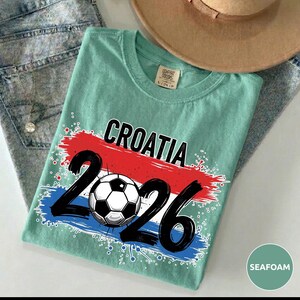 May include: Seafoam green t-shirt with the word "CROATIA" in black letters above a red and blue paint stroke design. Below the word "CROATIA" is the number "2026" in black with a soccer ball in the middle.