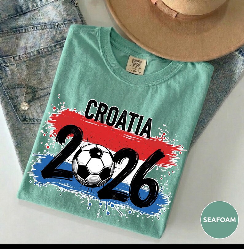 May include: Seafoam green t-shirt with the word "CROATIA" in black letters above a red and blue paint stroke design. Below the word "CROATIA" is the number "2026" in black with a soccer ball in the middle.