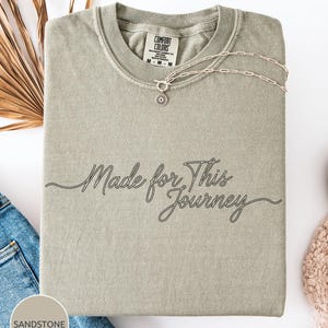 May include: A Sandstone-colored Comfort Colors t-shirt with the text "Made for This Journey" in a cursive font. A silver necklace with a flower pendant is draped across the shirt. The shirt is folded neatly on a white surface.