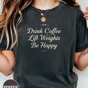 May include: Dark gray t-shirt with the text "Drink Coffee Lift Weights Be Happy" in a white, elegant font. The shirt has a crew neck and short sleeves. A small, round "Pepper" logo is visible at the bottom left.