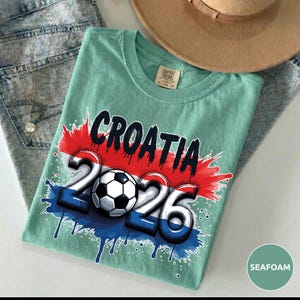 Może przedstawiać: Miętowa koszulka z napisem "CROATIA" w kolorze czarnym oraz liczbami "2026" z piłką nożną pośrodku. Projekt zawiera kolory chorwackiej flagi. Brązowy kapelusz i dżinsy znajdują się w tle.