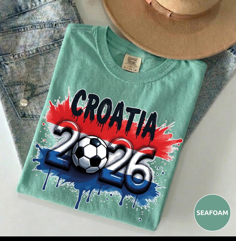 Może przedstawiać: Miętowa koszulka z napisem "CROATIA" w kolorze czarnym oraz liczbami "2026" z piłką nożną pośrodku. Projekt zawiera kolory chorwackiej flagi. Brązowy kapelusz i dżinsy znajdują się w tle.