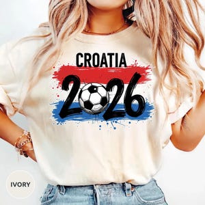 May include: Ivory-colored t-shirt featuring the word "CROATIA" above the numbers "2026" with a soccer ball in the center. The design includes red and blue brushstroke accents, and the shirt has a crew neck.