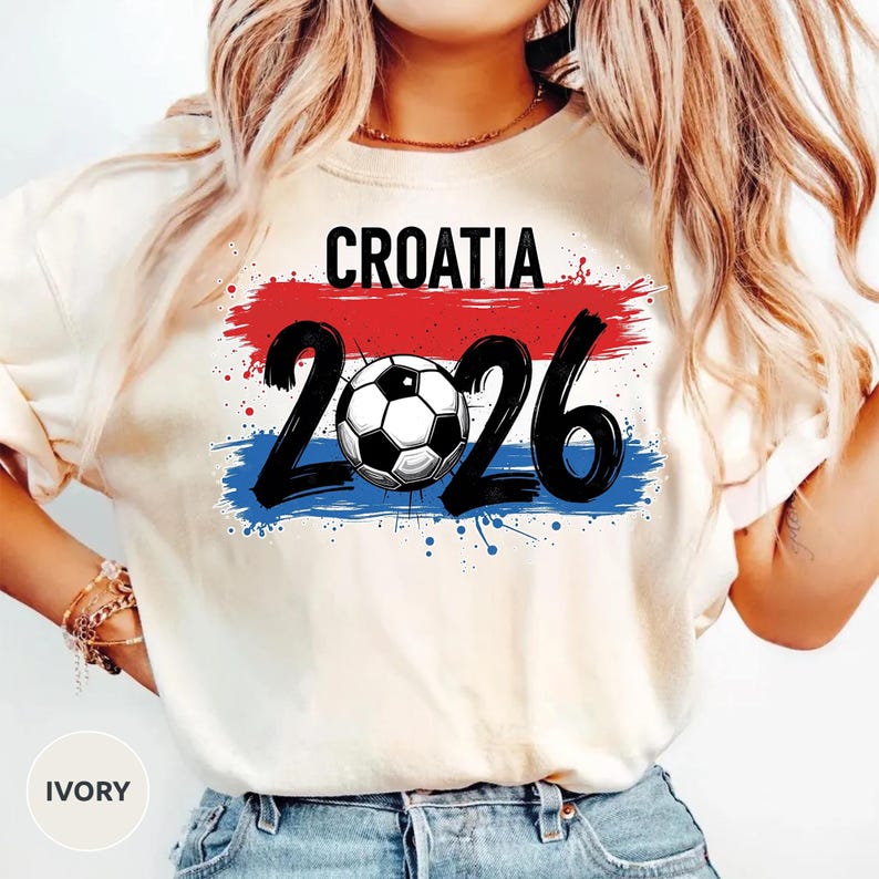 May include: Ivory-colored t-shirt featuring the word "CROATIA" above the numbers "2026" with a soccer ball in the center. The design includes red and blue brushstroke accents, and the shirt has a crew neck.