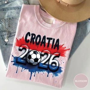Może przedstawiać: Jasnor&oacute;żowa koszulka z napisem "CROATIA" w kolorze czarnym i czerwonym oraz cyframi "2026" w kolorze niebieskim i białym, z piłką nożną pośrodku. Projekt ma efekt rozprysku z czerwoną i niebieską farbą. Beżowy kapelusz i niebieskie dżinsy są w tle.