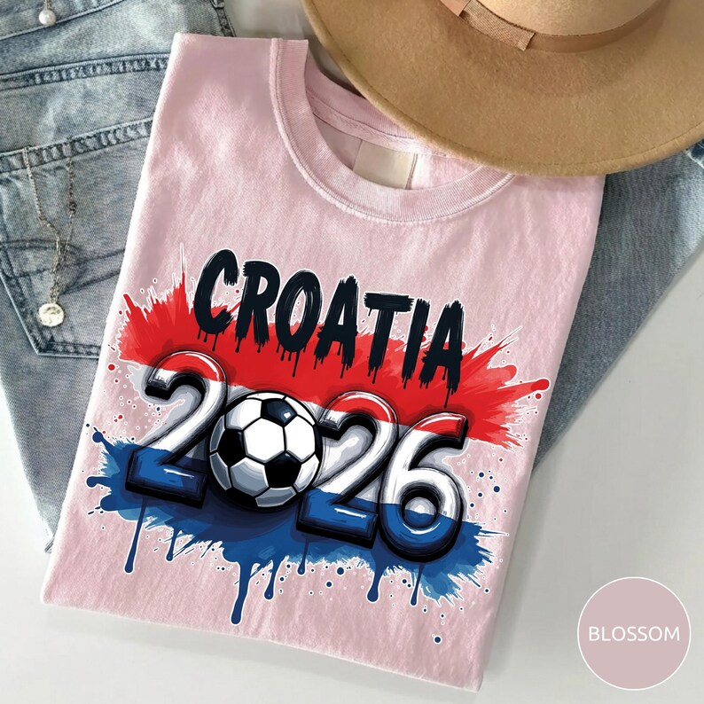 Może przedstawiać: Jasnor&oacute;żowa koszulka z napisem "CROATIA" w kolorze czarnym i czerwonym oraz cyframi "2026" w kolorze niebieskim i białym, z piłką nożną pośrodku. Projekt ma efekt rozprysku z czerwoną i niebieską farbą. Beżowy kapelusz i niebieskie dżinsy są w tle.