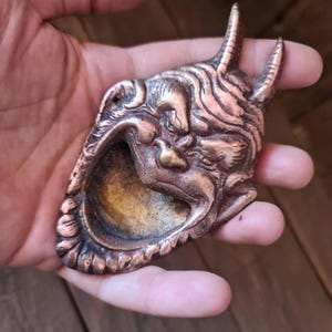 May include: A copper-coloured mask with a demonic face, featuring horns and an open mouth. The mask has intricate details and a weathered appearance, suggesting an antique or handcrafted item. The design evokes a sense of ancient artistry and cultural significance.