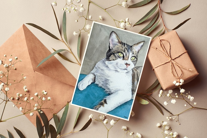Puede incluir: Una pintura de acuarela de un gato atigrado con ojos verdes, acostado sobre un coj&iacute;n azul turquesa. La pintura est&aacute; en una tarjeta blanca y rodeada de flores blancas y hojas verdes.