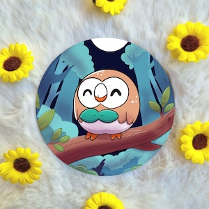 May include: A round sticker featuring a cartoon owl with brown and orange feathers, blue eyes, and a green leaf around its neck. The owl is perched on a branch in a forest setting with blue and green foliage.