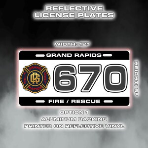 May include: A reflective license plate featuring "Grand Rapids" and the number "670". The plate measures 30.5 cm wide and 15.2 cm high, with a fire department emblem and the words "Fire / Rescue".