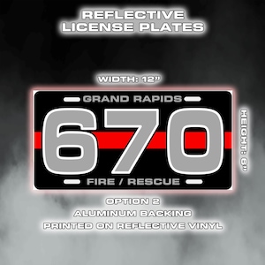 May include: Black license plate with the text "GRAND RAPIDS" and "FIRE / RESCUE". The plate features the number "670" in grey with a red stripe. The plate is 30.48 cm wide and 15.24 cm high. The text "REFLECTIVE LICENSE PLATES" is at the top.