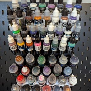 Könnte beinhalten: Eine Sammlung verschiedener Acrylfarbenflaschen und -töpfe, die auf einer schwarzen Lochwand angeordnet sind. Der obere Bereich zeigt zahlreiche Tropfflaschen mit weißen und schwarzen Verschlüssen, während der untere Bereich runde Farbtöpfe mit farbigen Deckeln enthält.