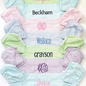 May include: A stack of six personalised baby bloomers in various colours with embroidered names and monograms. The bloomers are made of a soft, striped fabric.