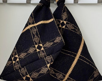May include: A black and tan plaid tote bag hanging on a white wall hook. The bag has a triangular shape with a repeating diamond pattern in tan and black. The bag is made of a soft, textured material.