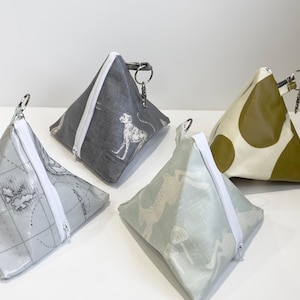 May include: Four pyramid-shaped pouches with zip closures and keyrings. Designs include a dog, a world map, a light green pattern, and olive green with white spots. The pouches are made of fabric and are pyramid-shaped.
