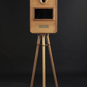 May include: A wooden photo booth with a rectangular body, a camera lens, and a screen. The booth is mounted on a tripod with leather accents. The wood has a natural grain and a warm, light brown color. The background is dark.
