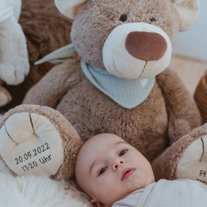 Könnte beinhalten: Ein großer, kuscheliger Teddybär mit hellbraunem Körper, brauner Nase und hellblauem Halstuch. Auf dem Pfoten des Bären sind Datum und Uhrzeit gestickt. Ein Baby liegt vor dem Bären.