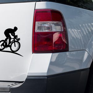 Puede incluir: Una silueta negra de un ciclista montando una bicicleta en un coche blanco. El ciclista lleva un casco y está en posición de carrera. El coche es un SUV blanco con una luz trasera roja.