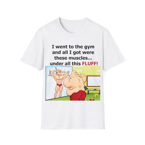 Pode incluir: Camiseta branca com uma ilustração humorística de um homem musculoso e um homem mais corpulento com o texto "Fui para a academia e tudo o que consegui foram esses músculos... sob todo esse FLUFF!"