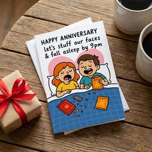 May include: A greeting card with the text "HAPPY ANNIVERSARY let's stuff our faces & fall asleep by 9pm" features an illustration of a couple in bed. A wrapped gift with a red bow and two mugs of coffee are on a wooden surface.