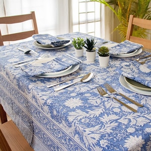 May include: A dining table set for four with a blue and white floral tablecloth, matching napkins, and white plates. Silverware and small potted plants complete the setting. Wooden chairs surround the table.