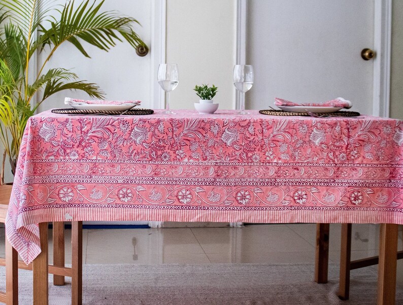 May include: A pink floral tablecloth with a white border and a repeating pattern of flowers and leaves. The tablecloth is laid on a wooden table with two place settings.