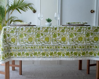 May include: A white tablecloth with a repeating pattern of green floral and leaf designs. The tablecloth is draped over a wooden table, with plates and glasses set on top. The overall theme is dining and home decor.