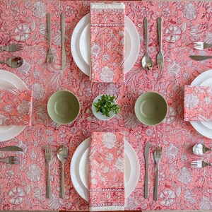 May include: A pink floral tablecloth with four place settings. Each place setting includes a white plate, a green bowl, a napkin, a fork, a knife, and a spoon. The napkins are folded and placed on top of the plates. The tablecloth has a white floral pattern on a pink background.