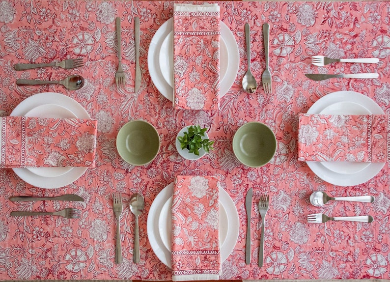 May include: A pink floral tablecloth with four place settings. Each place setting includes a white plate, a green bowl, a napkin, a fork, a knife, and a spoon. The napkins are folded and placed on top of the plates. The tablecloth has a white floral pattern on a pink background.