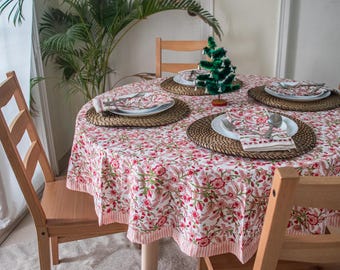 May include: A round table with a pink and white floral tablecloth. The table is set with four place settings, each with a white plate, a napkin, and silverware. There is a small green Christmas tree in the center of the table.