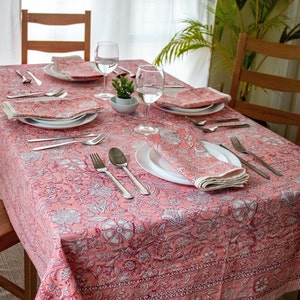 May include: A pink floral tablecloth with a white pattern set for a dinner party with plates, silverware, napkins, and glasses.