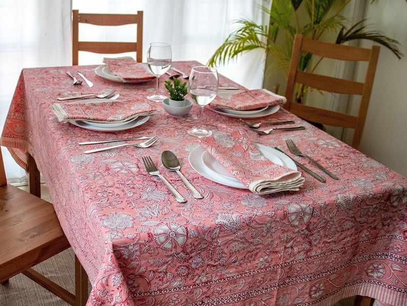 May include: A pink floral tablecloth with a white pattern set for a dinner party with plates, silverware, napkins, and glasses.