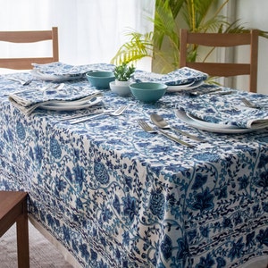 May include: A rectangular tablecloth with a blue floral pattern covers a wooden table set for a meal. White plates, blue bowls, and silverware are arranged on the table. Napkins with matching patterns are placed next to the plates.