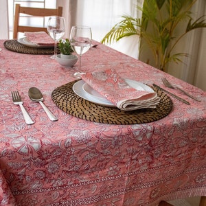 May include: A pink tablecloth with a floral pattern and a white border. The tablecloth is set with two place settings, each with a woven placemat, a white plate, a napkin, silverware, and a glass.