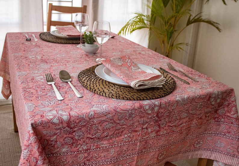 May include: A pink tablecloth with a floral pattern and a white border. The tablecloth is set with two place settings, each with a woven placemat, a white plate, a napkin, silverware, and a glass.