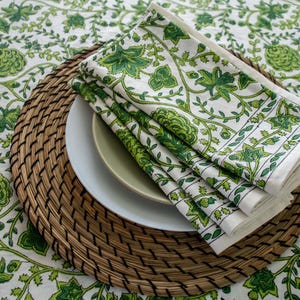May include: A set of white linen napkins with a green floral pattern. The napkins are folded and placed on a woven placemat with a white plate on top.