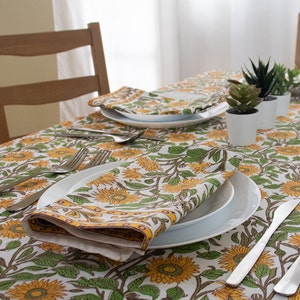 K&ouml;nnte beinhalten: Ein Esstisch mit einem floralen Sonnenblumenmuster. Der Tisch ist mit wei&szlig;en Tellern, Besteck und gefalteten Servietten gedeckt. Kleine Topfpflanzen sind auf dem Tisch platziert. Die Tischdecke und Servietten weisen ein gelbes, gr&uuml;nes und wei&szlig;es Sonnenblumenmuster auf.