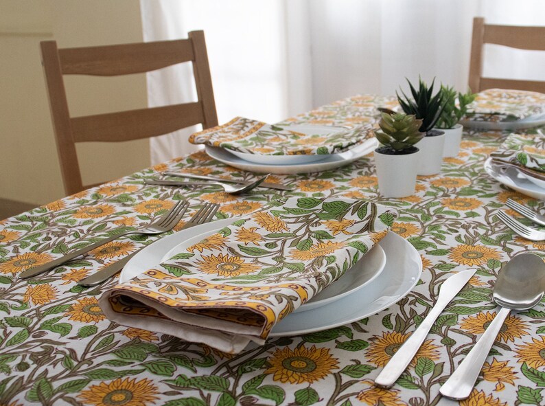 K&ouml;nnte beinhalten: Ein Esstisch mit einem floralen Sonnenblumenmuster. Der Tisch ist mit wei&szlig;en Tellern, Besteck und gefalteten Servietten gedeckt. Kleine Topfpflanzen sind auf dem Tisch platziert. Die Tischdecke und Servietten weisen ein gelbes, gr&uuml;nes und wei&szlig;es Sonnenblumenmuster auf.