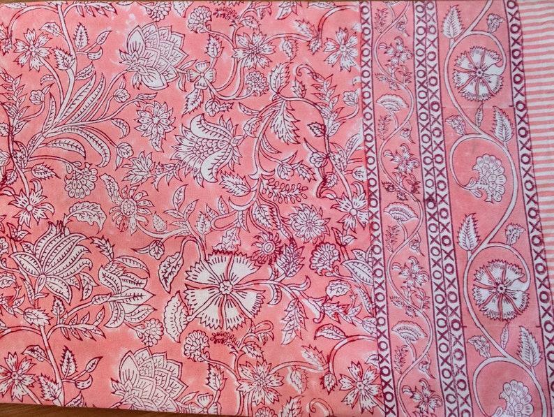 May include: A pink fabric with a white floral pattern. The fabric has a border with a repeating pattern of white circles and Xs.