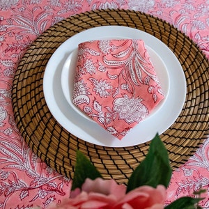 May include: A pink floral patterned napkin folded on a white plate, set on a woven placemat. The placemat is on a pink floral tablecloth.