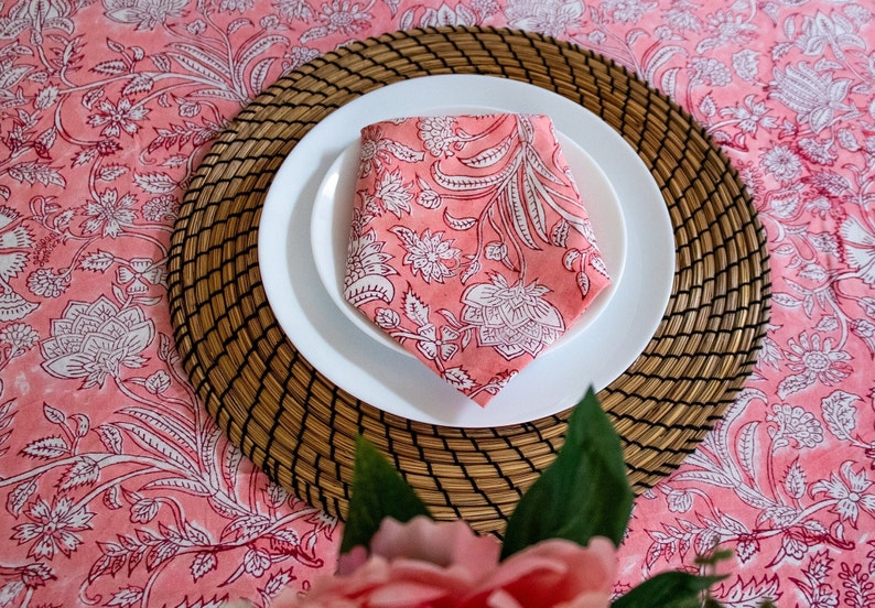 May include: A pink floral patterned napkin folded on a white plate, set on a woven placemat. The placemat is on a pink floral tablecloth.