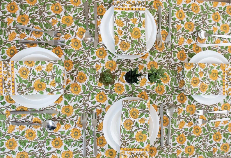 Mustard Floral Block-print cotton Tablecloth lay out on a table with a set of 4 napkins placed on white porcelain plates and cutlery beside each plate.