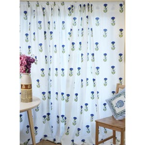 Peut inclure: Rideaux blancs avec un motif floral bleu répété. Les rideaux sont suspendus devant une fenêtre. Une petite table en bois contient un vase avec des fleurs. Une chaise en bois avec un oreiller est à droite.