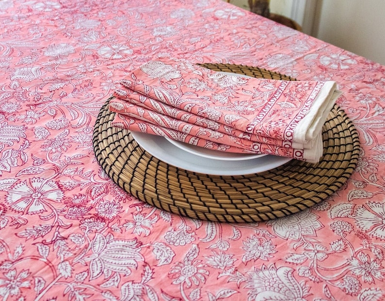 May include: Pink floral tablecloth with a woven placemat and folded pink floral napkins on top of a white plate.