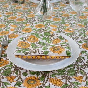 K&ouml;nnte beinhalten: Eine wei&szlig;e Tischdecke mit einem sich wiederholenden Muster aus gelben Sonnenblumen und gr&uuml;nen Bl&auml;ttern. Eine gefaltete Serviette mit dem gleichen Muster liegt auf einem wei&szlig;en Teller, mit Besteck darum angeordnet. Eine Vase und Gl&auml;ser stehen ebenfalls auf dem Tisch.