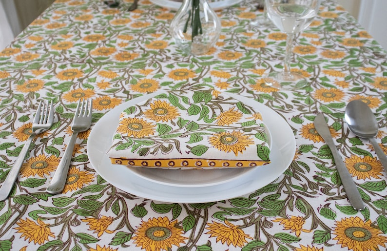 K&ouml;nnte beinhalten: Eine wei&szlig;e Tischdecke mit einem sich wiederholenden Muster aus gelben Sonnenblumen und gr&uuml;nen Bl&auml;ttern. Eine gefaltete Serviette mit dem gleichen Muster liegt auf einem wei&szlig;en Teller, mit Besteck darum angeordnet. Eine Vase und Gl&auml;ser stehen ebenfalls auf dem Tisch.