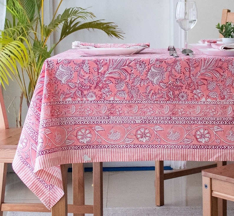 May include: A pink floral tablecloth with a white border and a repeating pattern of 'x's. The tablecloth is laid on a wooden table with two chairs.