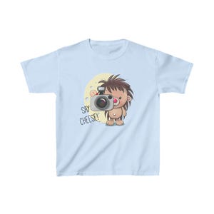 May include: Light blue t-shirt with a cartoon hedgehog holding a camera. The text "Say Cheese!" is printed on the shirt.